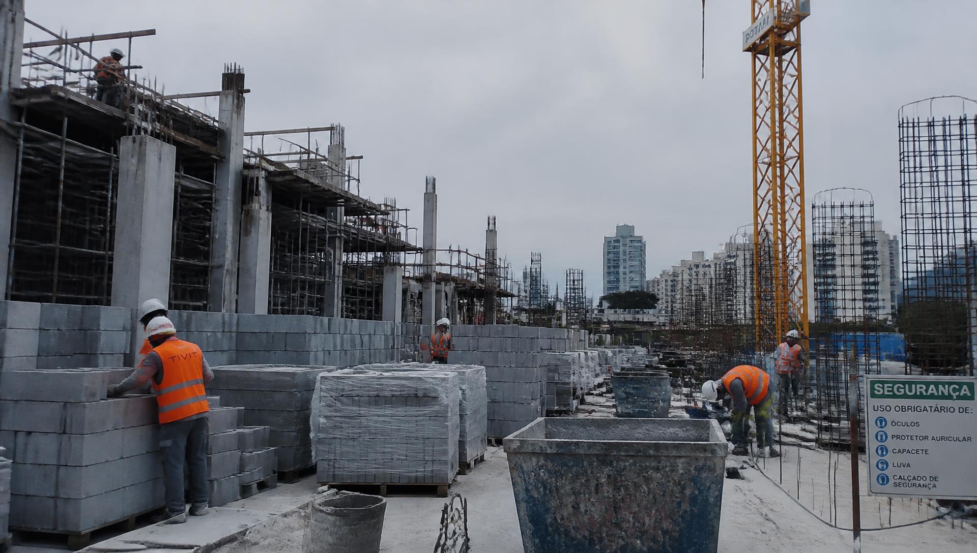 Equipe de obra executando alvenaria em canteiro com estrutura de concreto, materiais paletizados e grua ao fundo